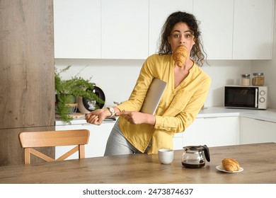 Hurrying young African-American woman with croissant and laptop in kitchen Foto stock