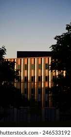 The image depicts a modern building at sunset, with the sun casting warm golden hues on its facade. Trees frame the building on both sides. Foto Stock
