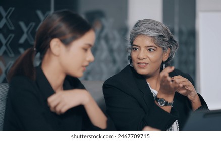 Indian business women sitting on chair using tablet look screen discuss share new project plan ideas job office place. Young adult colleagues old elder female doing work task together indoor workplace Stock-foto