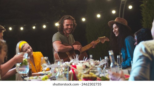 저녁에 정원에서 파티에서 즐거운 다양한 젊은 사람들. 기타를 연주하고 야외에서 노래를 부르는 행복한 백인 남성. 얼굴에 웃는 예쁜 흑인 소녀. 여가 개념 스톡 사진