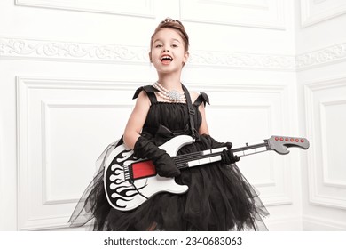 Little celebrity girl. A funny pretty girl in a luxury evening dress and with an evening hairstyle plays the electric guitar and sings. Pop and rock music culture. – Ảnh có sẵn