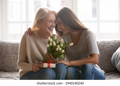 Loving young adult female child congratulate excited elderly mother with birthday anniversary at home. Smiling caring grownup millennial daughter present gift flowers to old mom on women s day. Stock Photo