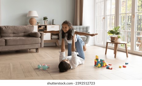 Loving young Indian mother raise on outstretched arms her cute little 6s daughter, family play together, dreaming about travel feel untroubled and happy. Leisure, games with children at home concept 库存照片