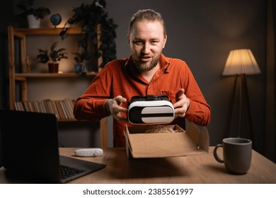 Man unpacking parcel with VR Glasses, looking inside, sitting at home desk, customer opening cardboard box with online store order and recording unboxing video, good shipping delivery service 库存照片