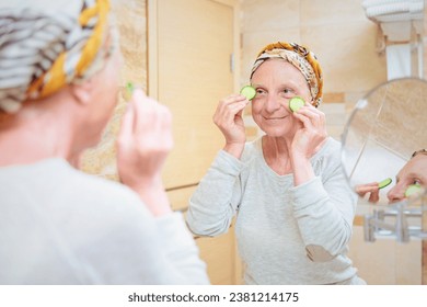 Mature woman taking care of her face. Senior woman standing at the mirror in the bathroom and applying cream or mask on her face and slices of cucumbers on her eyes 库存照片