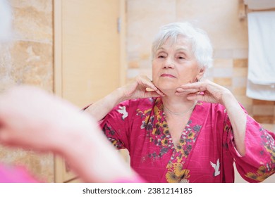 Mature woman taking care of her face. Senior woman standing at the mirror in the bathroom and applying cream to her face or doing self-massage 库存照片