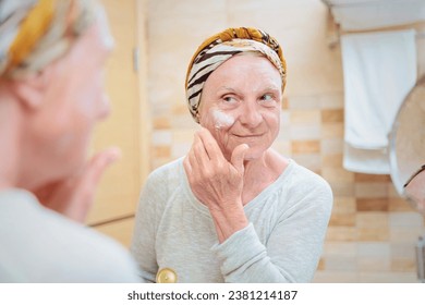 Mature woman taking care of her face. Senior woman standing at the mirror in the bathroom and applying cream or mask on her face 库存照片