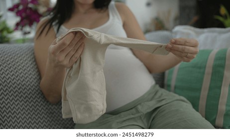 母親への期待 – 妊娠後期の妊娠中の母親は誇らしげにカメラに赤ちゃんの服を表示し、ソファに快適に座って、次の母性のための喜びと興奮を放射しますの写真素材