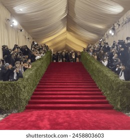 Met Gala red carpet scene, rich fabrics, red velvet carpet, gleaming golden light fixtures, excited crowd in elaborate outfits, antique furniture, grand staircase draped in lush green floral decorations, exotic orchids and elegant roses, elaborate crystal