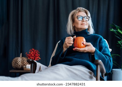 Middle aged woman relaxing with pumpkin shaped cup of hot drink in scandy style hygge interior home with fall mood decor. Lady dreaming, enjoy calm mood without stress, well being alone. Cozy autumn Foto Stock
