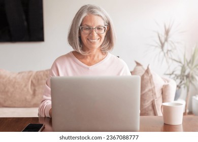 Middle aged mature woman using wireless laptop apps browsing internet sit at the table, smiling gray-haired lady working distantly on computer surfing web communicating online looking at screen at hom Foto stock