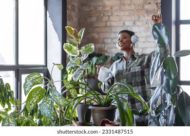 Morning routine and mediation, mental health concept. Cozy green interior. Attractive plus size African American young woman taking care about home plants. Enjoying household activity, singing, dance 库存照片
