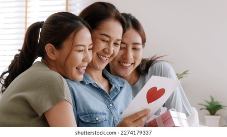 Mother day two grown up child cuddle hug give flower gift box red heart card to mature mum. Love kiss care mom asia middle age adult three people sitting at home sofa happy smile enjoy family time. Stock Photo