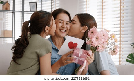 Mother day two grown up child cuddle hug give flower gift box red heart card to mature mum. Love kiss care mom asia middle age adult three people sitting at home sofa happy smile enjoy family time. Stock Photo