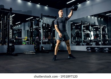 A muscular man in a gray T-shirt is doing a cross-fit workout in a gym with low lighting. He is in a squat position and does a dead lift with a kettle bell that he lifted over his head. Power boost Foto stock