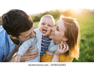 New parents holding small toddler, baby, outdoors in spring nature. Making baby laugh, kissing him on cheeks 库存照片