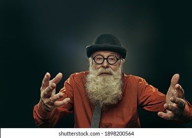 古風なボウリングハットと眼鏡をかけた老人が、訴える気配のしるしとして腕を横に広げた。 黒い背景。 の写真素材