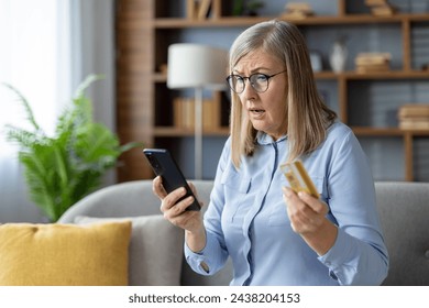 Una mujer mayor parece confundida mientras sostiene una tarjeta de crédito y mira su teléfono inteligente, posiblemente lidiando con compras en línea o desafíos tecnológicos. Foto de stock