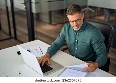 Older Indian business man, busy mature executive manager holding financial accounting papers checking banking income documents file report using laptop computer working in office sitting at desk., fotografie de stoc
