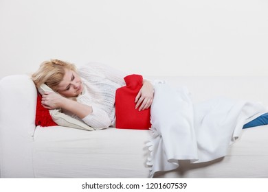 Painful periods and menstrual cramp problems concept. Woman having stomach cramps lying on cofa feeling very unwell holding hot water bottle to feel some relief Foto de stock