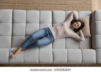 Peaceful young woman in casual clothes relaxing alone on cozy sofa with hands behind head and eyes closed, above view. Fatigue relief, comfortable furniture store ad, carefree leisure at home concept Foto Stock