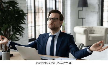 Peaceful business leader meditating at workplace, making zen hands and fingers at laptop. Calm businessman relaxing in office, breathing, doing yoga, practicing mindfulness and stress relief exercises Foto Stock