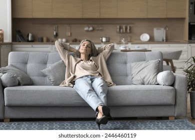 平和な中年の女性リビングルームで居心地の良いソファに座って、頭の後ろに手を傾けて、怠惰な週末を楽しんで、モダンな家で昼寝を楽しんでいます。疲労軽減、休憩、リラクゼーションの写真素材