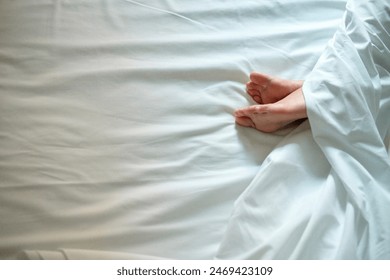 A person's bare feet protrude from under a white duvet on a bed with white linens. Arkistovalokuva