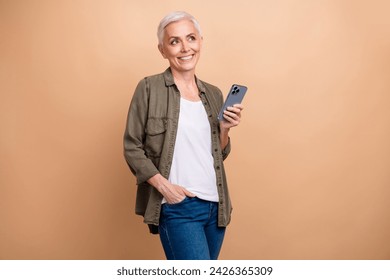 Photo of positive grey hair businesswoman wear khaki shirt using cellphone look empty space count budget isolated on beige color background Stock fotografie