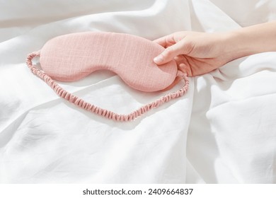 Pink eye mask for sleep on bed background, minimal lifestyle aesthetic scene, copy space. Top view woman hand holding sleeping mask on white bed linen, morning daylight. Comfort rest, healthy trend Foto Stock