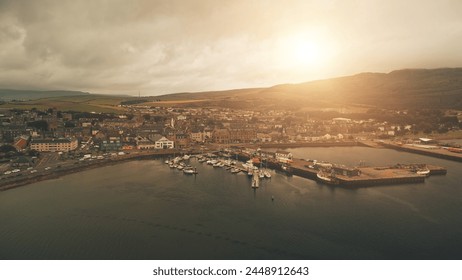 Pier town citycsape with historic buildings at ocean bay aerial. Sun shine over ships, yachts at marina wharf. Streets, road with cars, trucks. Green hill, valley at Campbeltown city, Scotland, Europe Stock Photo