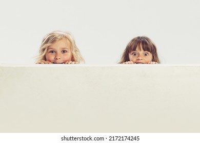 Portrait of two little beautiful girls, children peeking out the table isolated over grey studio background. Hide-and-seek. Concept of childhood, friendship, family, fun, lifestyle, retro fashion Foto stock