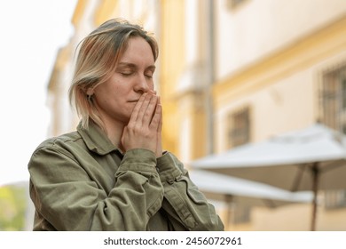 Portrait of young woman praying with closed eyes to God asking for blessing, help, forgiveness outdoors. Caucasian girl clasping hands wishing luck on sunny urban city street. Town religion lifestyles: stockfoto