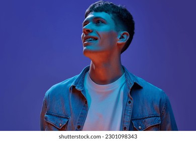 Portrait of young handsome guy in jeans shirt and white t-shirt design with smile, looking away against gradient purple background in neon light. Concept of human emotions, lifestyle, youth Foto stock