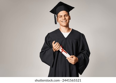 Стоковая фотография: portrait of young student in graduate gown and cap holding to diploma with hands on grey background