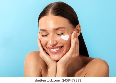 Portrait of cheerful beautiful lady with moisturizer cream in heart shape on her pretty face, recommend new beauty product, blue background: stockfoto