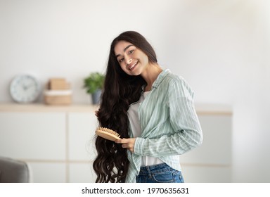 Portrait of lovely Indian lady brushing her wavy long hair, using wooden brush indoors. Cheerful young woman taking care of herself at home. Domestic spa salon, wellness and beauty concept Stock Photo
