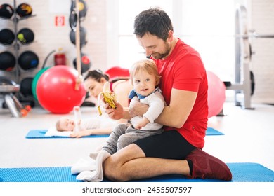 Portrait of new dad on group exercise class in gym, feeding baby with banana. Parents staying active while boding with babies. 库存照片