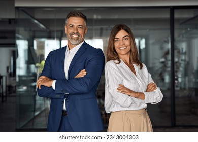 Portrait of smiling mature Latin or Indian business man and European business woman standing arms crossed in office. Two diverse colleagues, group team of confident professional business people. Foto stock