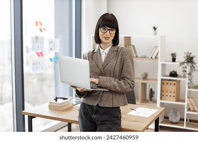 Стоковая фотография: Portrait of smiling caucasian adult businesswoman sitting on edge of wooden desk with laptop. Cheerful woman in formal wear managing successful company from modern workplace.