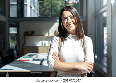 Portrait of smiling young brunette woman looking at camera standing with arms crossed in front of desk in coworking space. Female entrepreneur. Copyspace. Startup concept. Co working space concept. स्टॉक फ़ोटो