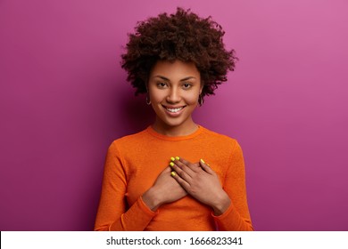 Pleased girl in orange jumper presses palms to heart, makes thankful gesture, touched with cordial congratulations, smiles positively, isolated over purple background. Acknowledgement concept Arkistovalokuva