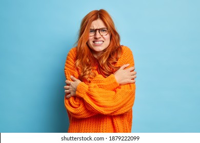 Redhead woman trembles from cold hugs herself to warm up walks outdoor during freezing temperature clenches teeth and shakes wears only sweater poses against blue background. Its chilly here Stockfoto
