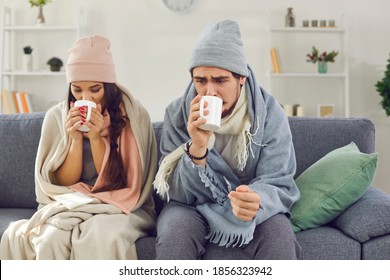 Sad sick young couple wrapped in plaid sitting on sofa in living-room drinking hot tea trying to warm up. Freezing man and woman suffering from cold or flu fever or having trouble with central heating स्टॉक फ़ोटो