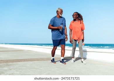 A senior couple joyfully walking together along a sunny beachside promenade, sharing laughter and good times in casual sporty attire. – Ảnh có sẵn