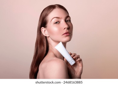 Стоковая фотография: sensitive skin care concept, beautiful red-haired young woman with freckles holds a white cream tube mock up in her hand, no makeup natural beauty isolated on beige background, clean glowing skin