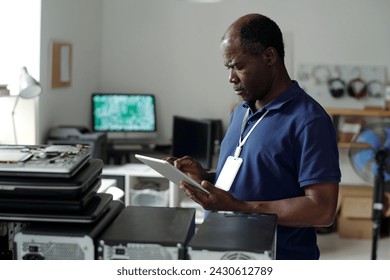 職場でカメラの前に立ち、オンラインマニュアルを通して見ているタブレットを持つ深刻な成熟したアフリカ系アメリカ人の技術者の写真素材