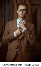 Serious young man in a smart brown three-piece suit and spectacles adjusts his tie on the background of luxury classic suits in a premium men's clothing store. Men's business fashion. 库存照片