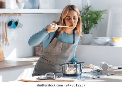 Shot of beautiful woman cooking healthy food in casserole while blowing the spoon to taste the food in the kitchen at home. Foto stock