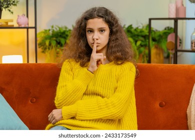 Shh be quiet please. Child girl presses index finger to lips makes silence hush gesture sign do not tell gossip secret. Teenager female 14-15 years kid at home apartment living room sits on couch Foto Stock